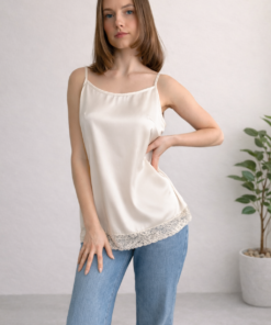 Hodvábna košieľka vo farbe ivory s tenkými ramienkami a čipkovým lemom na modelke v džínsoch s rukou na boku.